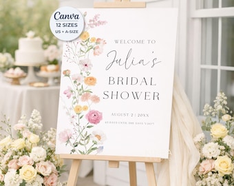May include: A white rectangular bridal shower welcome sign on a wooden easel, featuring a vertical floral arrangement of pink, yellow, and white flowers. The text reads: Welcome to Julia's Bridal Shower, August 2 | 20XX, 63 days until she says I do.