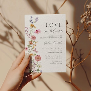 May include: A floral wedding invitation with the text "LOVE in bloom" and details for the event. The invitation features a watercolor floral design in pink, purple, and yellow. The invitation is held in a hand.