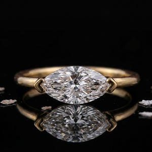 May include: A gold ring featuring a marquise-cut diamond. The diamond is set in a gold band with a simple, elegant design. The ring is displayed on a reflective black surface, highlighting its brilliance.