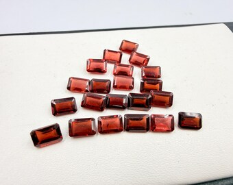 May include: A collection of rectangular, faceted, red-orange gemstones. The gems are arranged on a white surface, showcasing their clarity and color. The gemstones are likely for jewelry making.