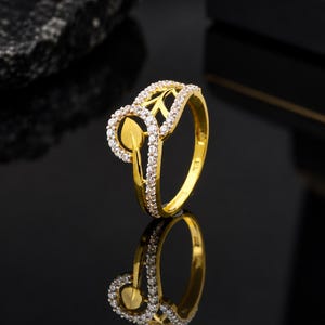 May include: A gold ring featuring a leaf-shaped design element accented with small, clear, round-cut stones. The band is polished gold, and the decorative top section is lined with a row of sparkling stones that follow the curved, organic shape.