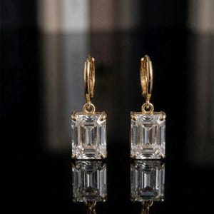 May include: A pair of gold-coloured hoop earrings featuring rectangular, clear gemstones. The earrings are set against a dark backdrop, with their reflections below. The gemstones are emerald-cut.