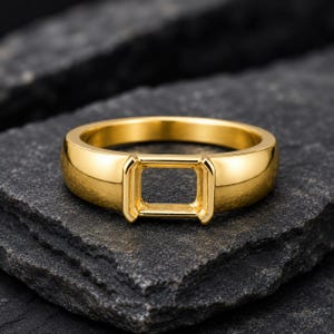 Op de afbeelding: Een gepolijste goudkleurige ring met een gladde, ronde band en een opvallende, lege rechthoekige zetting in het midden, ontworpen om een edelsteen te bevatten. Het metalen oppervlak heeft een reflecterende, glanzende afwerking met randen.