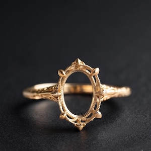 Könnte beinhalten: Ein goldener Ringrohling mit ovalem Kopf, aufwendigen Milgrain-Details und sechs Krappen. Die Ringschiene zeigt an den Seiten kunstvolle, texturierte Metallarbeiten, die zu einer zentralen, offenen Fassung für einen Edelstein führen.