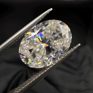 Op de afbeelding: Een ovaal geslepen diamant, vastgehouden door een pincet tegen een zwarte achtergrond. De diamant schittert met een regenboog aan kleuren, waarbij de facetten en de schittering worden getoond. De pincet is zilverkleurig en heeft een getextureerde grip.