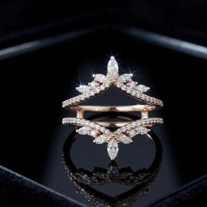 Può includere: Set di anelli in oro rosa con un design scintillante. Gli anelli presentano una serie di piccole pietre chiare rotonde e a forma di marquise. Il design è posto su uno sfondo scuro.