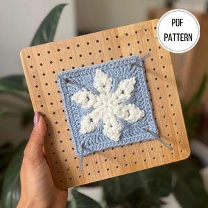 Può includere: Un quadrato di fiocco di neve all'uncinetto azzurro e bianco è appuntato a un blocco di legno. Il disegno del fiocco di neve è centrato, con il testo "PDF PATTERN" in un cerchio bianco in alto a destra.