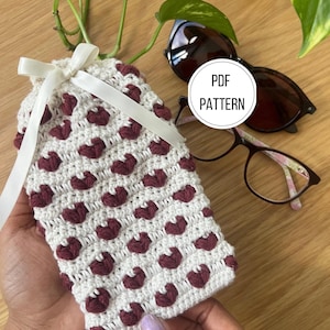 May include: A handmade crochet glasses case with a white base and burgundy heart-shaped accents. The case is tied with a cream-coloured ribbon. The image also shows sunglasses and eyeglasses. The text "PDF PATTERN" is visible.
