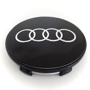 May include: A black wheel center cap with a silver Audi logo. The logo consists of four interlocking rings. The cap is round and has a glossy finish. The cap has two black clips on the bottom.