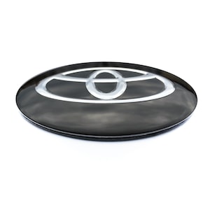 May include: A black oval car emblem with a silver and white Toyota logo. The emblem has a glossy finish and is set against a white background. The Toyota logo is a stylized design of overlapping ovals.
