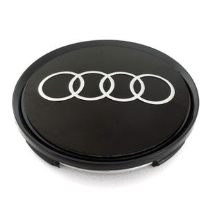 May include: A black wheel center cap featuring the Audi logo. The logo consists of four interlocking silver rings. The cap has a glossy finish and a black outer ring.