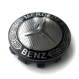 May include: A black and silver Mercedes-Benz emblem. The emblem features the Mercedes-Benz logo, a three-pointed star, in silver, surrounded by a black ring with the words "Mercedes" and "Benz" in white.