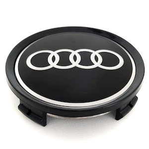 May include: Black and silver hubcap featuring the Audi logo, a set of four interlocking rings. The logo is white against a black background, surrounded by a silver trim. The hubcap has a black plastic base with securing clips.