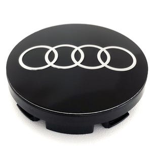 May include: A black hubcap with the Audi logo in white. The logo consists of four interlocking rings. The hubcap is round with a glossy finish and has several black tabs on the back.