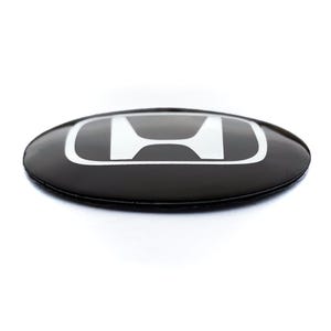 May include: A black and white Honda emblem with a glossy finish. The emblem is oval-shaped and features the Honda logo in white against a black background. The emblem is likely for a car or vehicle.