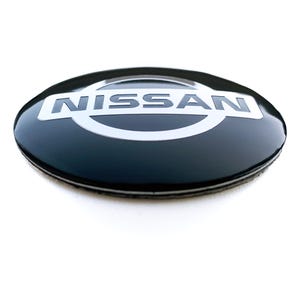 May include: A black and silver Nissan emblem. The oval-shaped emblem features the brand name "NISSAN" in silver block letters, set against a black background. The emblem has a glossy finish and is set against a white background.
