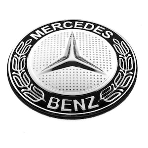 May include: A silver and black Mercedes-Benz emblem. The emblem features a three-pointed star in the center, surrounded by a silver circle with the word "Mercedes" at the top and "Benz" at the bottom. The emblem is framed by a black border.