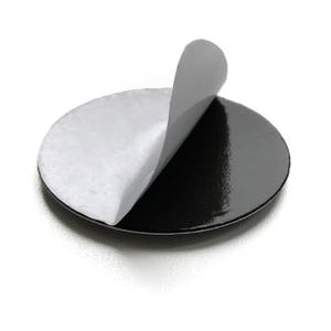May include: A circular adhesive pad with a glossy black finish on one side and a white protective paper backing partially peeled away from the top, revealing the sticky surface underneath. The disc is flat and uniform in its circular shape.