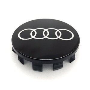 May include: A black wheel center cap with the Audi logo in white. The logo consists of four interlocking rings. The cap has a glossy finish and a series of tabs around the edge for secure attachment.