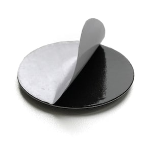 Pode incluir: Um autocolante preto e circular com um acabamento brilhante. O papel de suporte branco está parcialmente descolado, revelando o adesivo. O autocolante está sobre um fundo branco.