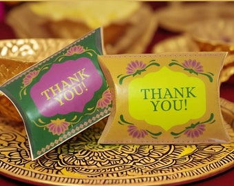 Puede incluir: Dos pequeñas cajas de almohada con diseños florales dorados, verdes y morados. Las cajas tienen "Thank You!" escrito en blanco.
