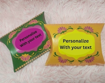 Puede incluir: Dos cajas decorativas en forma de almohada, una verde y otra amarilla, con diseños florales y el texto "Personalize With your text". Las cajas tienen forma de almohada y están decoradas con motivos florales rosas y dorados.