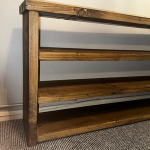 May include: A handcrafted wooden shelf with a rich, dark brown stain. The shelf has four horizontal levels supported by vertical side pieces. The wood grain is visible, adding to the rustic aesthetic. Perfect for storage or display.