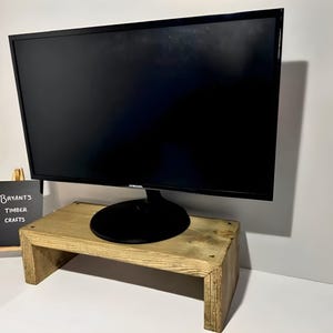 May include: A wooden monitor stand supporting a black computer screen. The stand is constructed from light-toned wood and is rectangular. A small chalkboard sign, reading "Bryant's Timber Crafts", is positioned to the left of the stand.