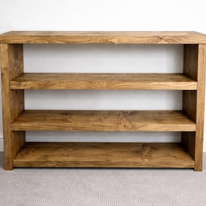 May include: A wooden bookshelf with three shelves. The shelves and frame are made of stained wood, showcasing the natural grain. The bookshelf is a warm brown color and stands against a white wall.