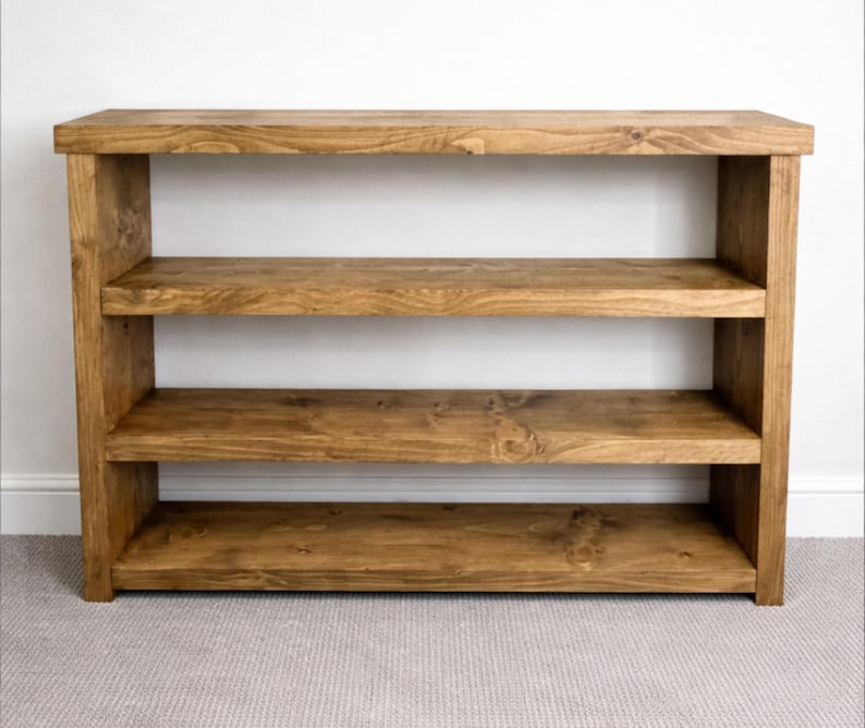 May include: A wooden bookshelf with three shelves. The shelves and frame are made of stained wood, showcasing the natural grain. The bookshelf is a warm brown color and stands against a white wall.