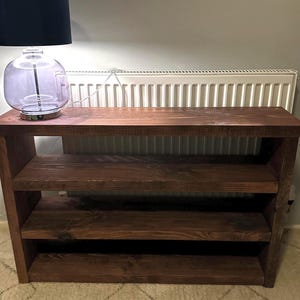 May include: A dark brown wooden shelf unit with three shelves. The unit has a rectangular top and sides, with the shelves supported by the sides. A lamp with a black shade and a clear glass base sits on top of the shelf.