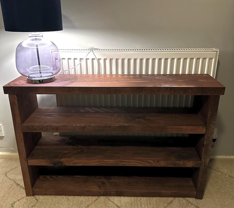 May include: A dark brown wooden shelf unit with three shelves. The unit has a rectangular top and sides, with the shelves supported by the sides. A lamp with a black shade and a clear glass base sits on top of the shelf.