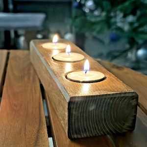 May include: A wooden candle holder with four candle slots, each with a lit tea light. The holder is made of dark brown wood with a natural grain pattern. The holder is sitting on a wooden table.