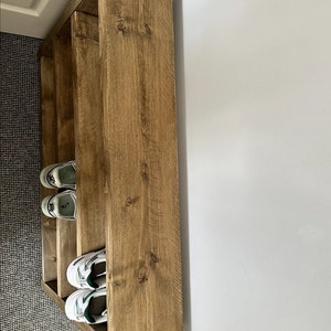 May include: A wooden shoe rack with three shelves. The rack is made of light brown wood and is positioned against a white wall. Several pairs of white and green sneakers are stored on the shelves.