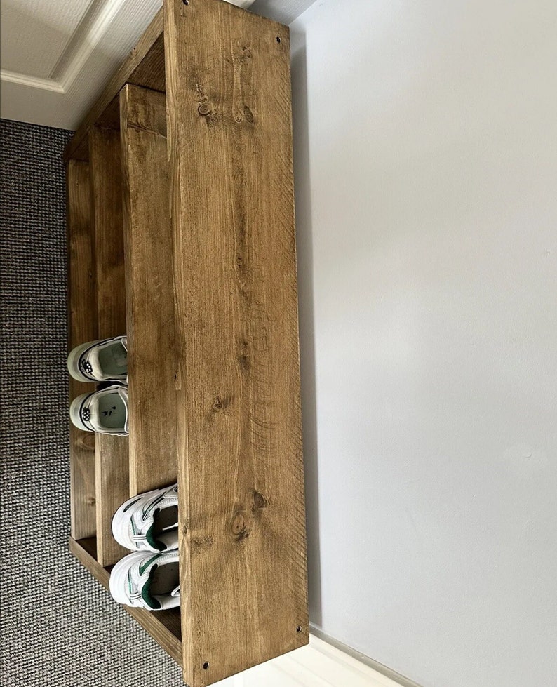May include: A wooden shoe rack with three shelves. The rack is made of light brown wood and is positioned against a white wall. Several pairs of white and green sneakers are stored on the shelves.