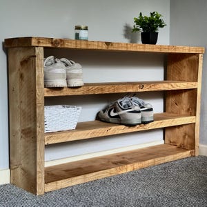 May include: A rustic, three-tiered wooden shoe rack with visible wood grain. The top shelf holds a small potted plant and a jar. Two pairs of shoes and a white basket are displayed on the lower shelves. The rack is made of light brown wood.