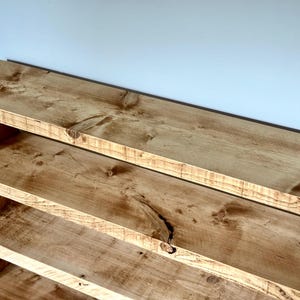 May include: A rustic wooden bookshelf with three shelves. The shelves and frame are made of light brown, knotty wood. The bookshelf is against a white wall.