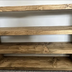 May include: A wooden bookshelf with three shelves. The shelves and frame are made of stained wood, showcasing the natural grain. The bookshelf is a warm brown color and is against a white wall.