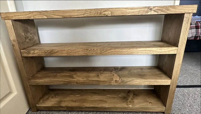 May include: A wooden bookshelf with three shelves. The shelves and frame are made of stained wood, showcasing the natural grain. The bookshelf is a warm brown color and is against a white wall.