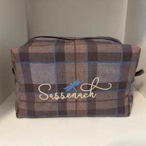 May include: A brown and blue plaid travel bag with the embroidered word "Sassenach" and a blue dragonfly design. The bag has a rectangular shape and a handle on each side. The bag is sitting on a white shelf.