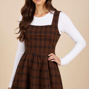 May include: A brown and black checked pinafore dress with a white long-sleeved shirt underneath. The dress has a square neckline and a flared skirt.