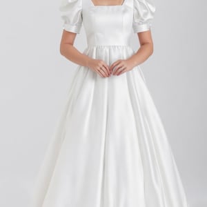 May include: A white satin wedding dress with a square neckline and puffed sleeves. The dress has a fitted bodice and a full skirt.