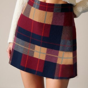 May include: A plaid mini skirt in a classic pattern of burgundy, navy, tan, and light blue. The skirt is worn with a cream-colored sweater and brown boots. The skirt's design features a traditional tartan pattern.