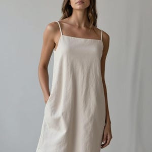 Può includere: Un abito color crema senza maniche con scollo quadrato e spalline sottili. L'abito ha una vestibilità ampia e arriva a metà coscia. Presenta due tasche anteriori ed è realizzato in un tessuto strutturato. La modella indossa l'abito.