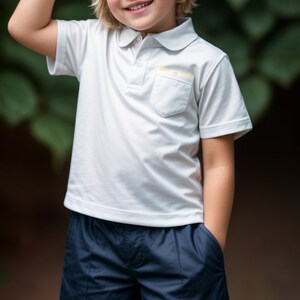 Puede incluir: Un ni&ntilde;o peque&ntilde;o con una camisa polo blanca con un peque&ntilde;o bolsillo en el pecho izquierdo y pantalones cortos azul marino. El ni&ntilde;o sonr&iacute;e y tiene el pelo rubio.
