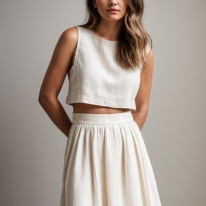 May include: A cream-colored, sleeveless crop top and matching midi skirt. The top has a round neckline, and the skirt is pleated. The fabric appears to be a textured linen or similar material. The outfit is a neutral, minimalist style.