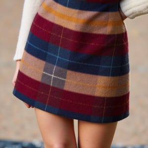 May include: A plaid mini skirt in shades of burgundy, navy blue, tan, and gold. The skirt is worn with a cream-colored sweater and brown boots. The skirt features a classic plaid pattern.