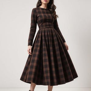Op de afbeelding: Een bruin en zwart geruite midi-jurk met lange mouwen en een getailleerd bovenlijf. De jurk heeft een volle rok met plooien en een ronde hals.
