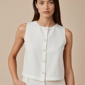 Könnte beinhalten: Ein ärmelloses, cremefarbenes Oberteil mit rundem Ausschnitt und einer Reihe von fünf passenden Knöpfen auf der Vorderseite. Das Oberteil wird mit einer weißen Hose kombiniert. Das Outfit ist schlicht und elegant, passend für verschiedene Anlässe.