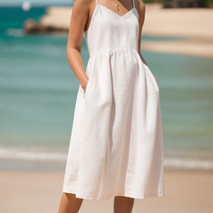 Puede incluir: Un vestido midi de lino blanco con tirantes finos y escote en V. El vestido tiene bolsillos a los lados y una cintura fruncida. La modelo lleva el vestido con sandalias negras y está de pie en una playa de arena.